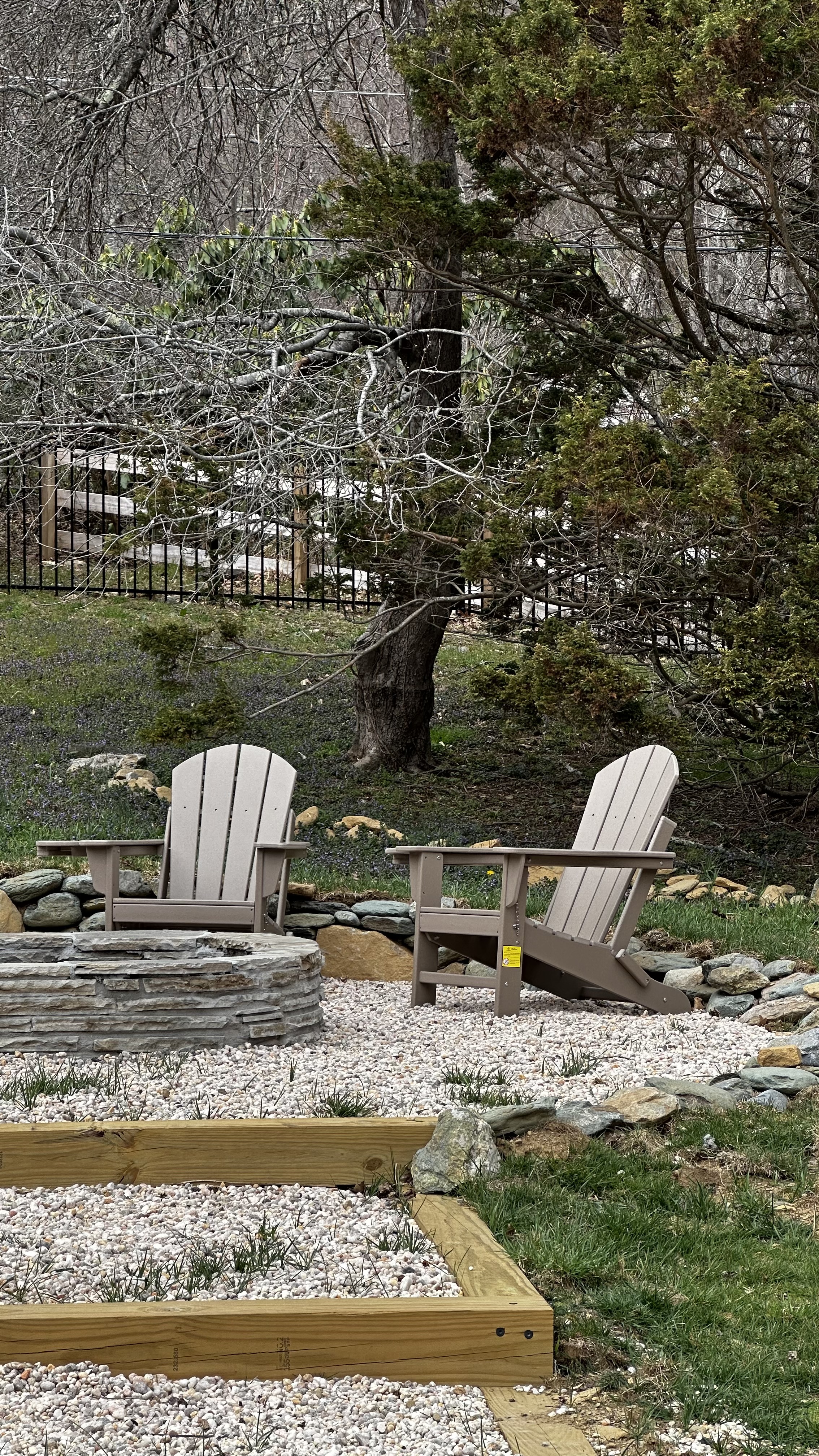 Two Adirondack chairs on a gravel patio sitting in front of a large rock.