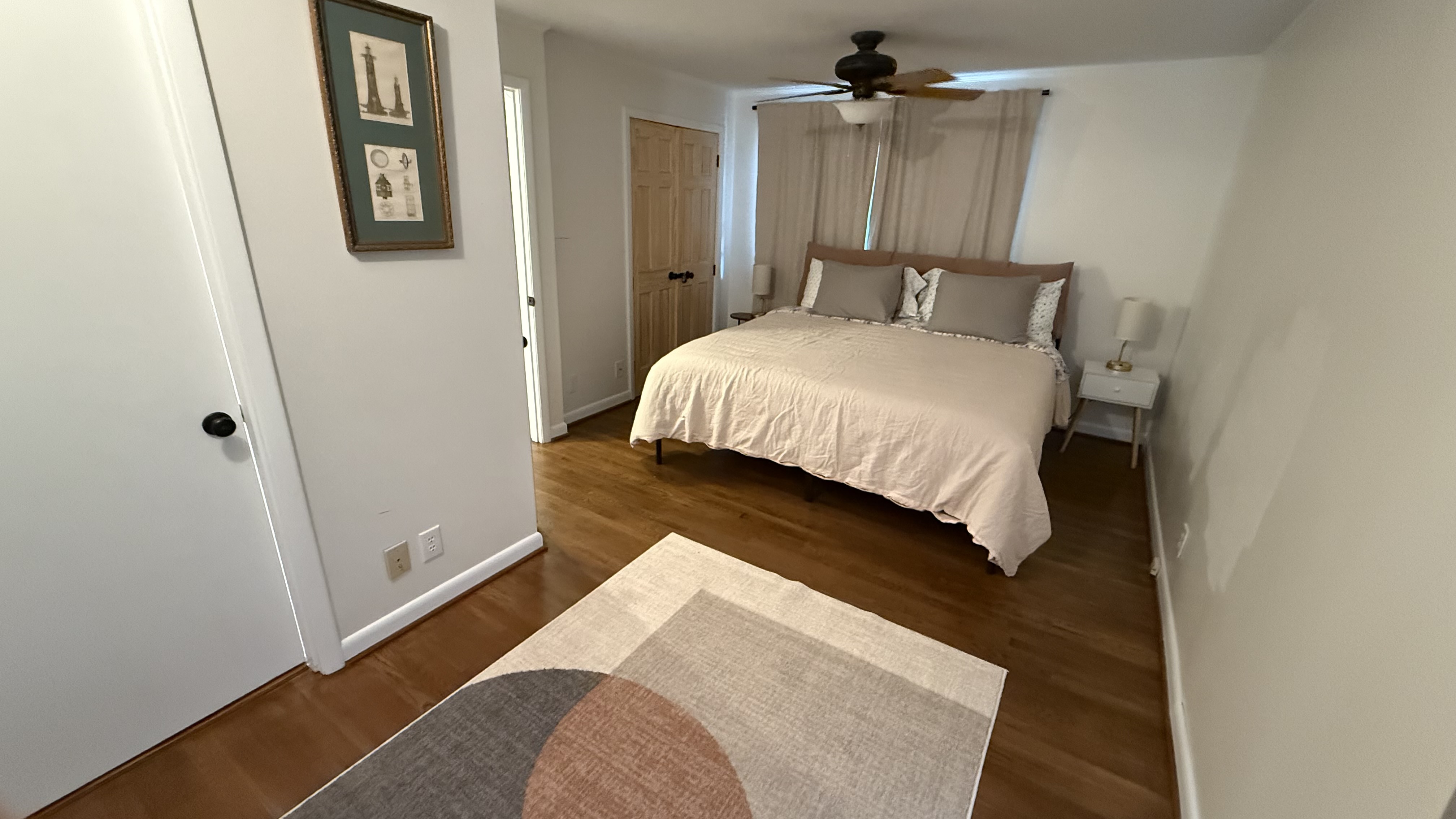 A small bedroom with an unmade bed in the rear of the room, a ceiling fan, with a bedside table to the right of the bed. A...