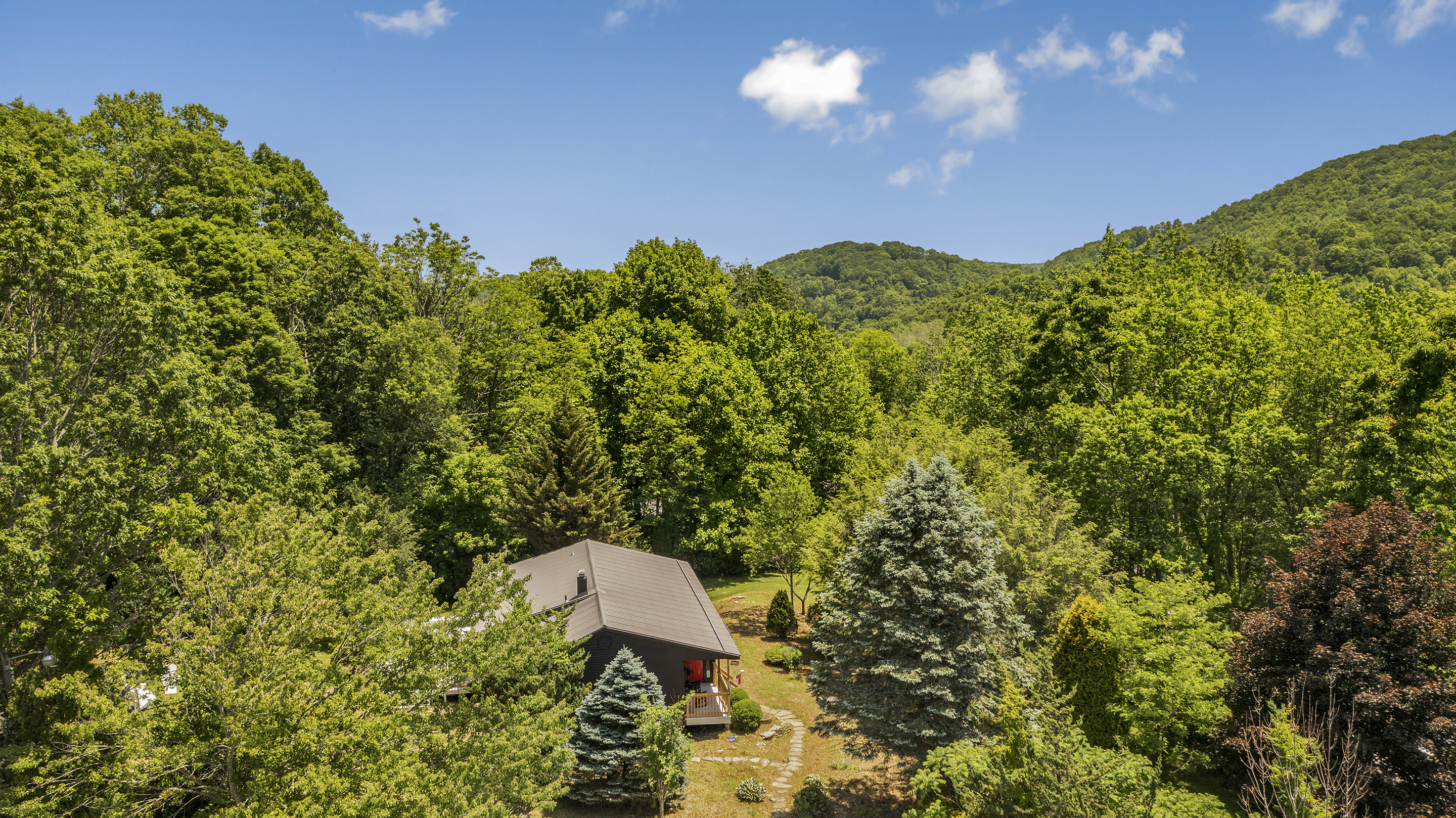 A picturesque house nestled in a vibrant, densely wooded area surrounded by trees and rolling hills under a clear, sunny d...